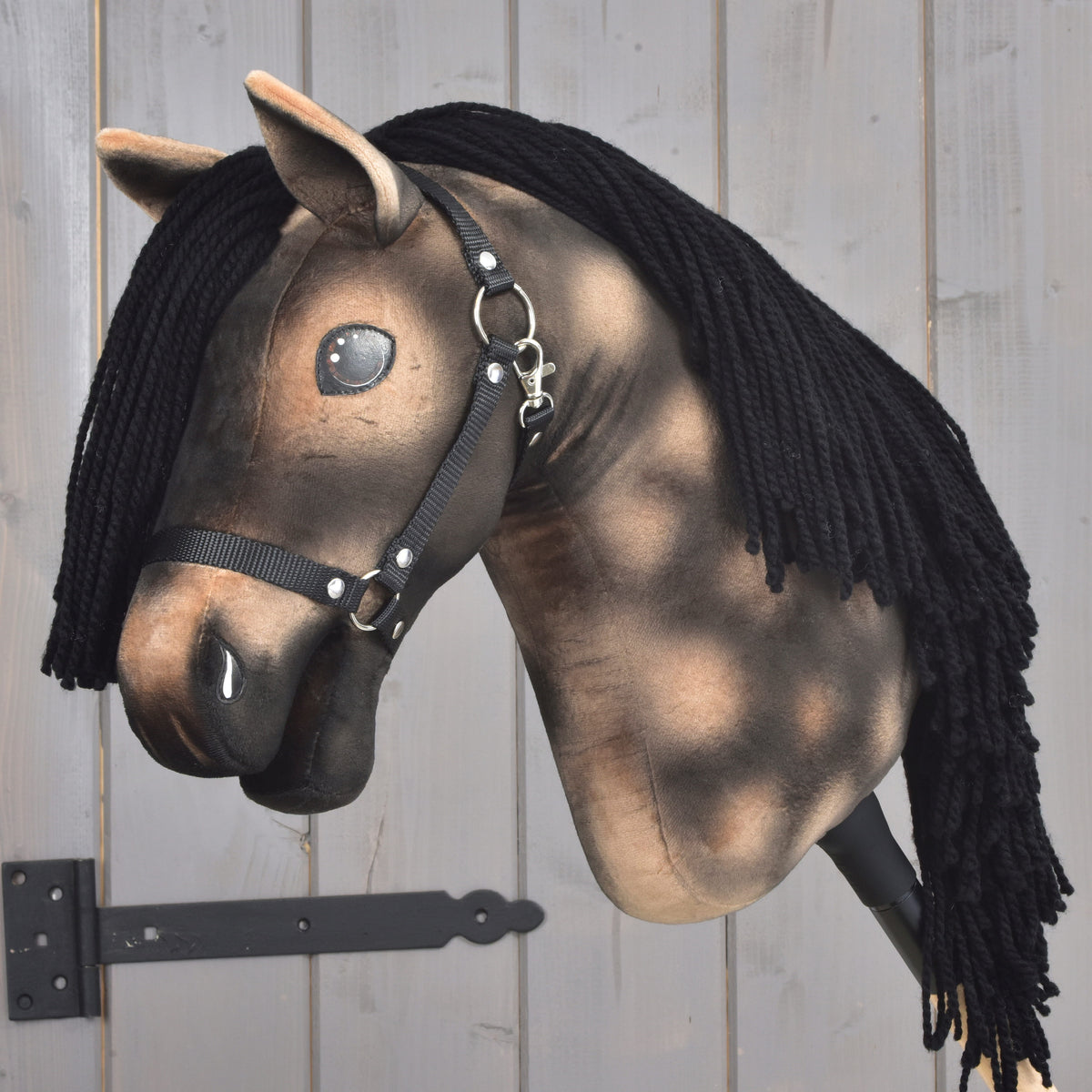 The Hobby Horse LarDen “Stormy” features a brown plush head, a black yarn mane, a black halter, and shiny button eyes mounted on a wooden stick against a light wooden background—an enchanting piece inspired by the spirit of a Hanoverian Stallion.