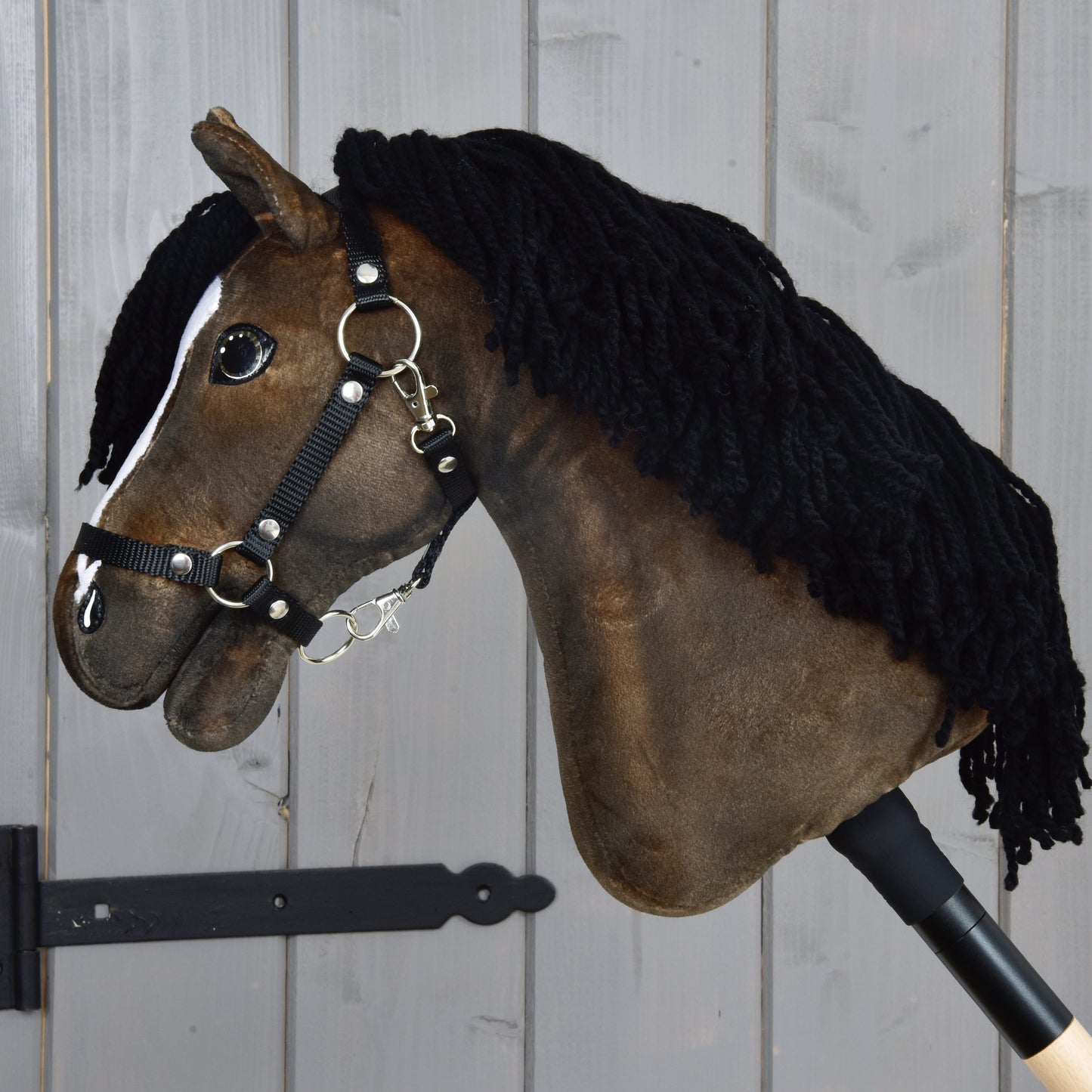 The Hobby Horse LarDen "Hobby Horse Sundance with Black Halter" features a brown body, black yarn mane, white facial stripe, black halter, and comes mounted on a black stick against a gray wooden background.