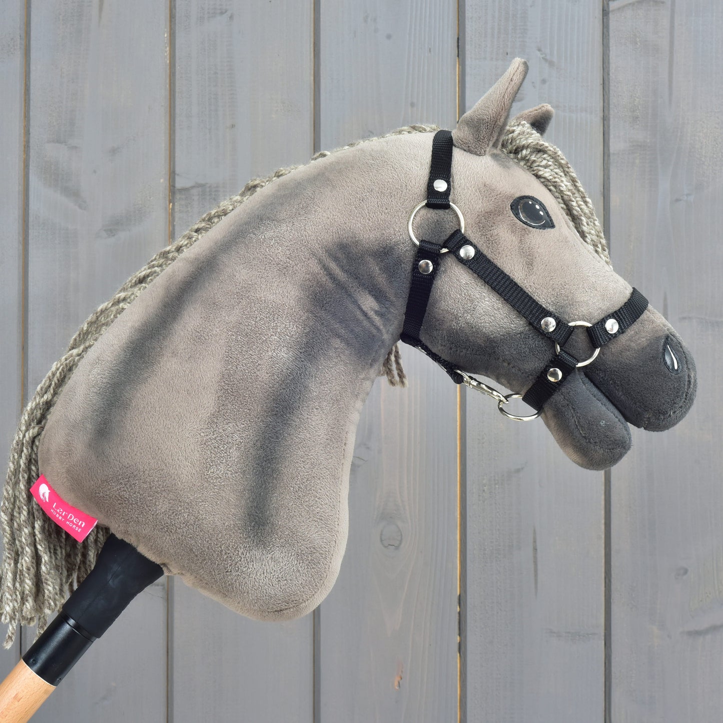 The Hobby Horse LarDen Hobby Horse Swing with black halter, size S, featuring a plush design with a grey mane and mounted on a wooden stick to capture the stately charm of a Hanoverian stallion, is displayed against a weathered grey wooden background.