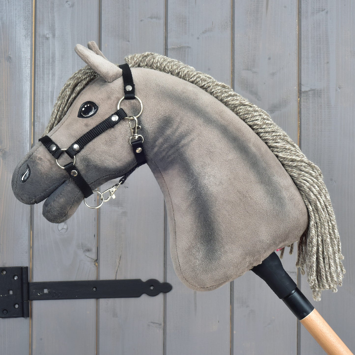 A gray plush hobby horse reminiscent of a Hanoverian stallion, featuring a black halter and rope mane, is mounted on a wooden stick and displayed in front of a wooden wall with visible grain and a black metal hinge, adding an air of elegance to the charming Hobby Horse Swing with black halter, size S by Hobby Horse LarDen.