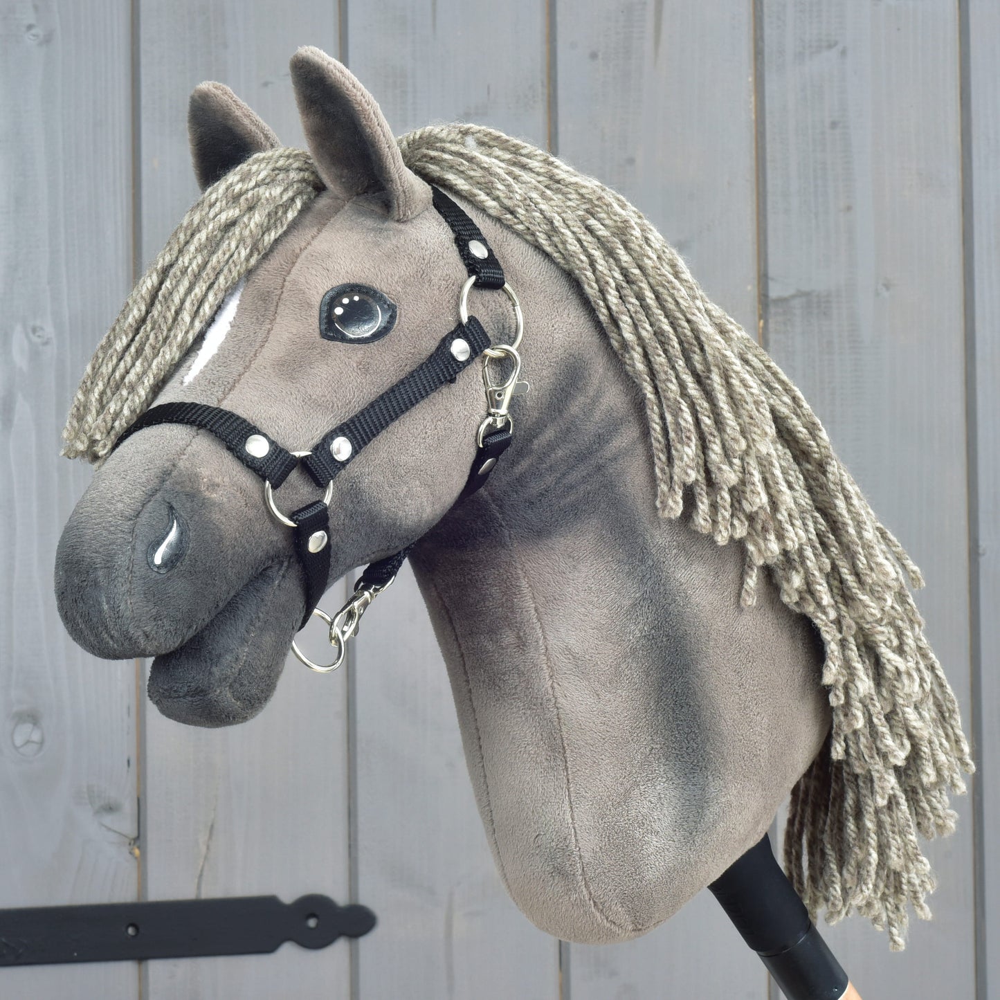 The Hobby Horse LarDen "Hobby Horse Swing with black halter," size S, features a plush horse with a braided yarn mane and a sleek black halter, mounted on a stick and showcased against a wooden backdrop.
