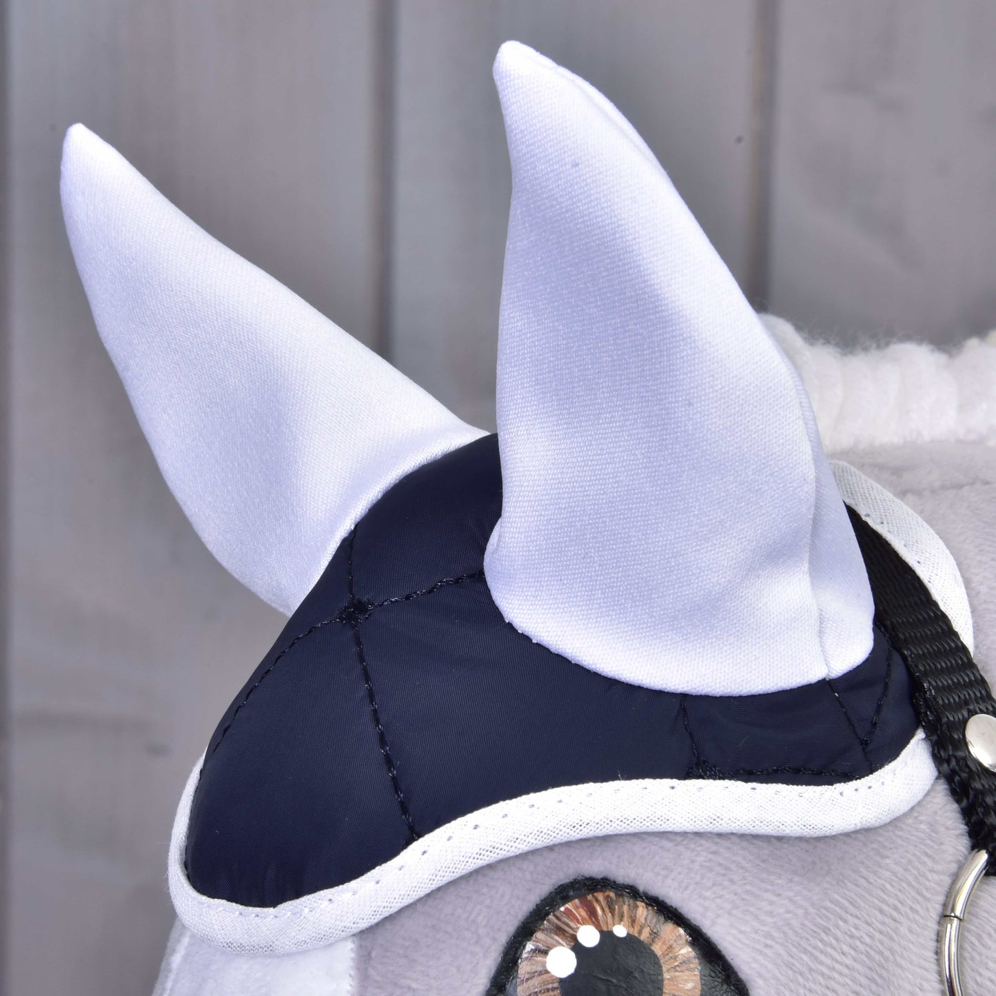 Gros plan sur une tête de cheval en peluche portant l’élégant Ensemble Thermo Bleu de Hobby Horse LarDen, avec oreilles blanches, bonnet bleu marine et blanc, et bride noire. Idéal comme élément décoratif devant des panneaux en bois clair.