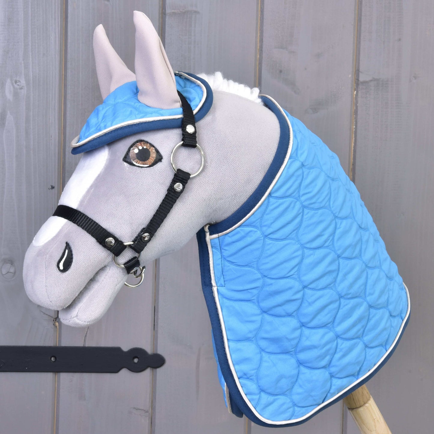 Le Hobby Horse en peluche gris de Hobby Horse LarDen porte la couverture de l’Ensemble Thermo Indigo et le bonnet d’oreilles assorti avec un filet, présenté comme une décoration de cheval devant un mur en bois.