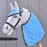 Das graue Plüsch-Hobby Horse von Hobby Horse LarDen trägt die Decke und das passende Fliegenhauben-Set Set Thermo Indigo mit Zaumzeug, dargestellt als charmante Dekoration vor einer Holzwand.