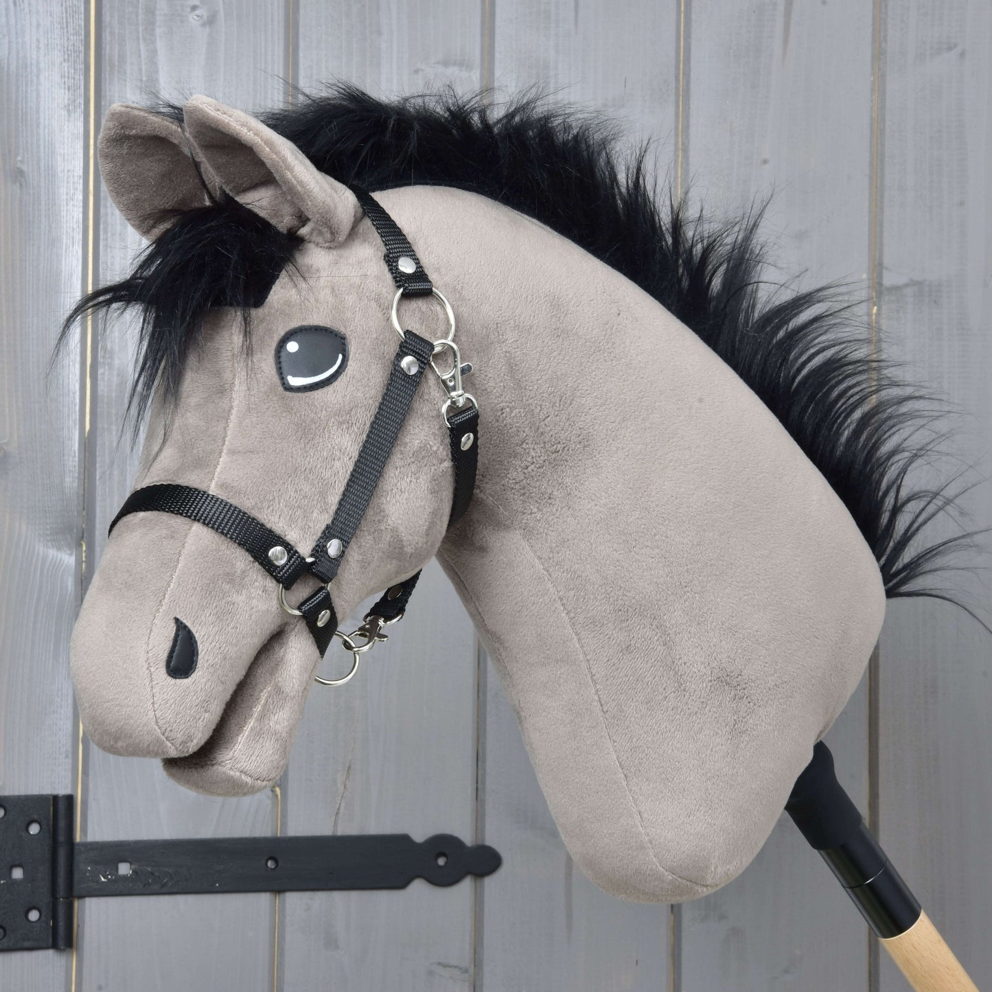 Hobby Horse Thor mit Schwarzem Halfter von Hobby Horse LarDen, ein Hobby Horse Hengst im Hannoveraner-Stil aus Plüsch, mit schwarzer Mähne, schwarzem Halfter und gestickten Augen, auf einem Holzstab vor einer hellgrauen Holzwand.