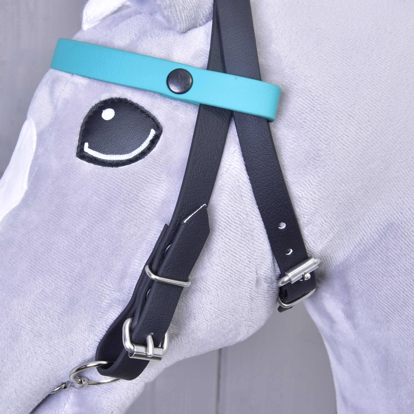 Close-up of Hobby Horse LarDen's Bridle with 2 headbands turquoise (Size M) on a light gray plush horse with a cartoon-style embroidered eye. The bridle features silver buckles and an interchangeable browband, no noseband, in teal and black.