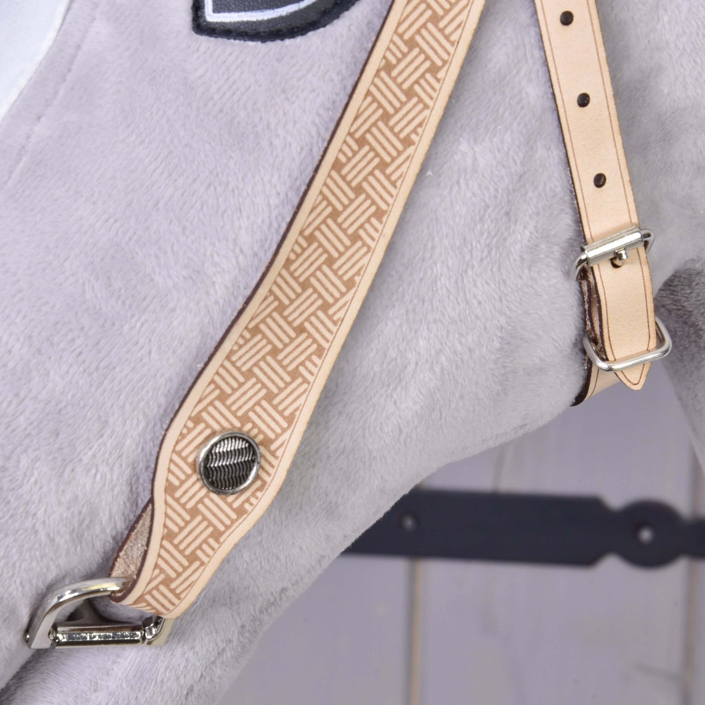 Gros plan sur la Bride Western Classique de Hobby Horse LarDen : sangle fauve en cuir premium avec motif géométrique et bouton métallique, fixée sur une surface en peluche gris clair, mettant en valeur un savoir-faire d’inspiration western sur fond flou.