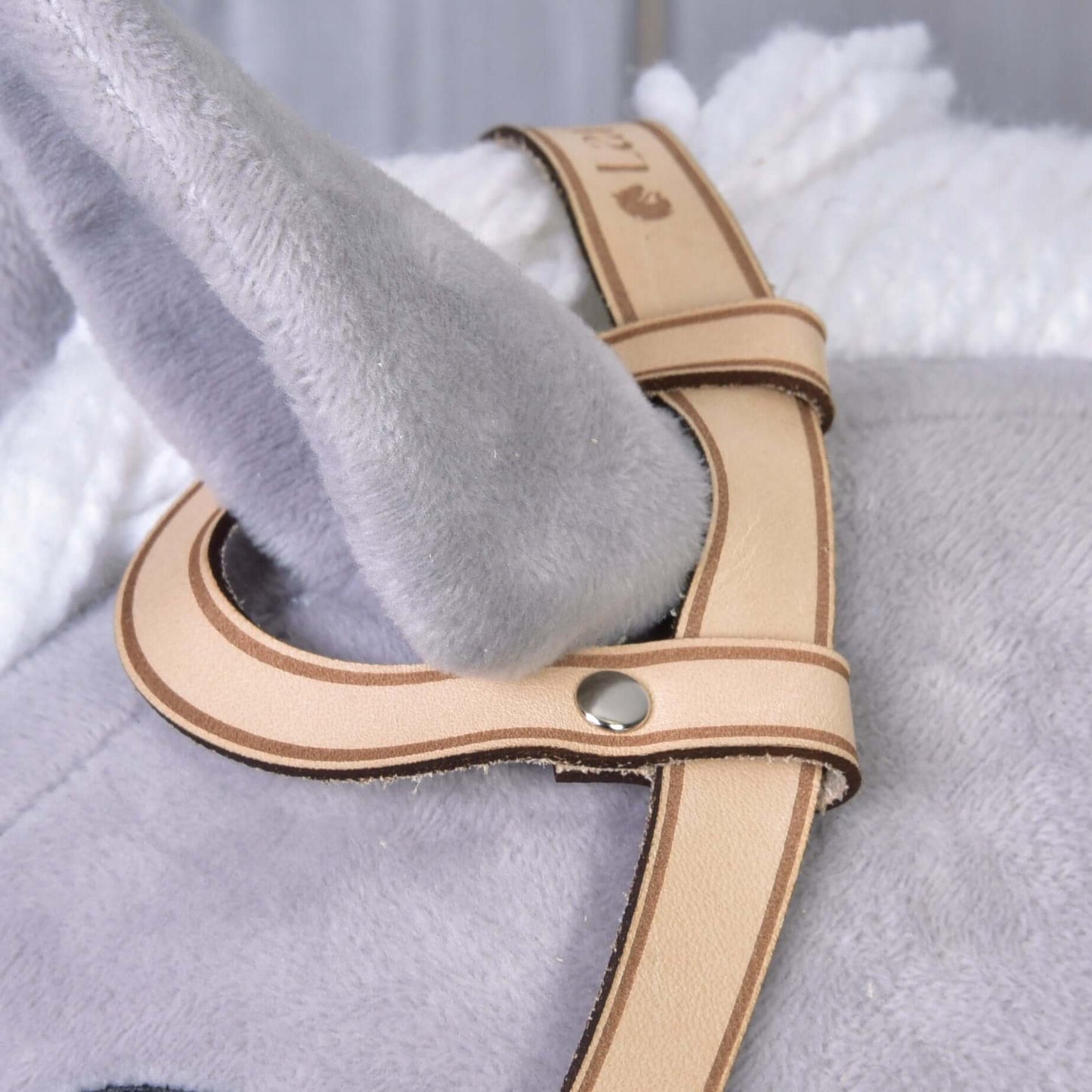 Nahaufnahme von grauem Plüschstoff und einer weichen weißen Decke mit Westerntrense Einohr von Hobby Horse LarDen – ein hellbrauner Riemen aus hochwertigem Leder mit Metallnieten, gelegt um ein weiches graues Stofftier-Ohr.
