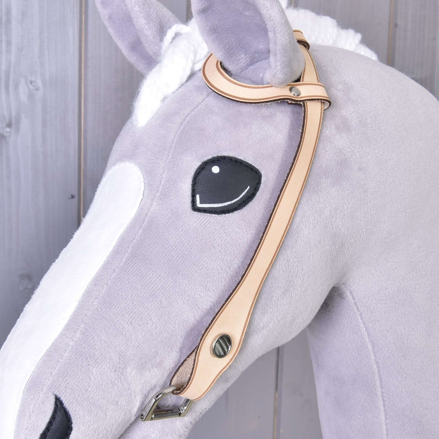 Nahaufnahme eines Plüschpferdekopfes aus hellgrauem und weißem Stoff mit aufgesticktem schwarzem Auge und Westerntrense Einohr von Hobby Horse LarDen mit silberfarbenen Beschlägen vor Holz-Hintergrund.