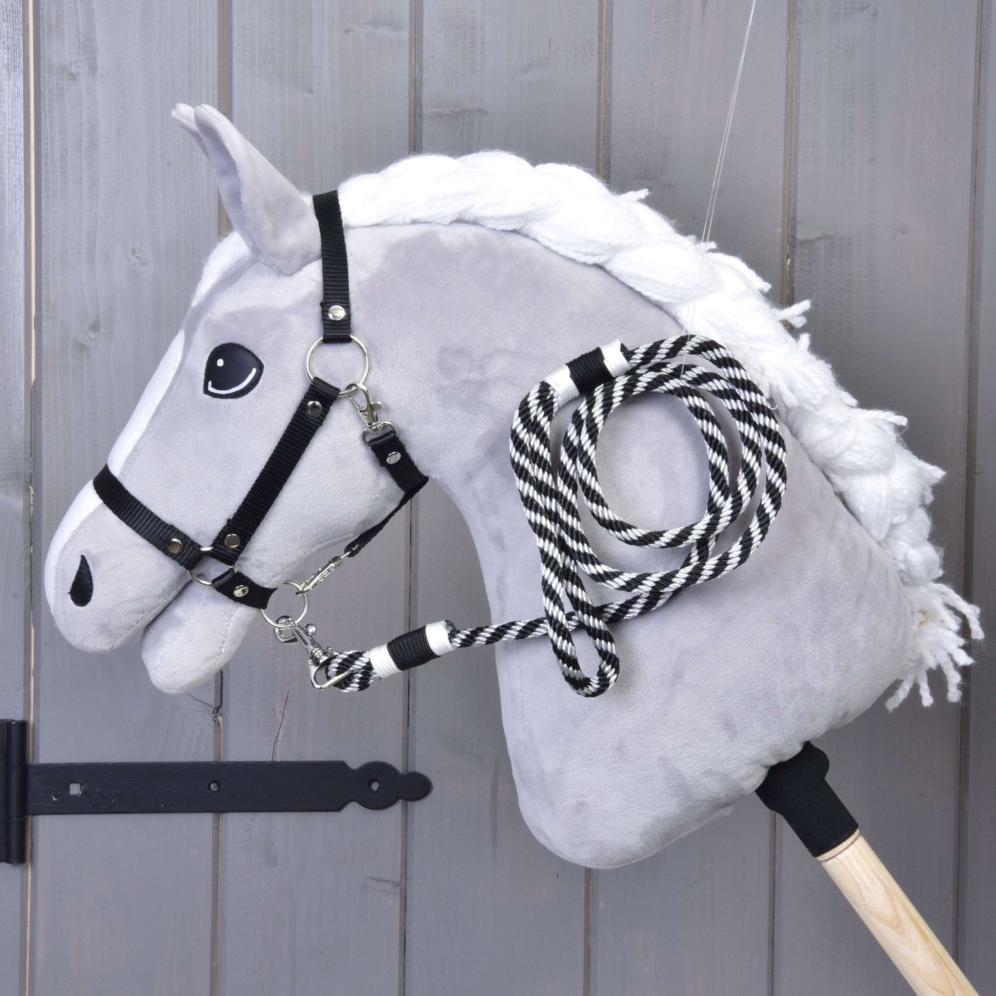 Hobby Horse - Longe rayée noire de Hobby Horse LarDen sur un cheval en peluche avec crinière blanche, bride noire et longe rayée noir et blanc, monté sur un bâton en bois et présenté devant un mur en bois.