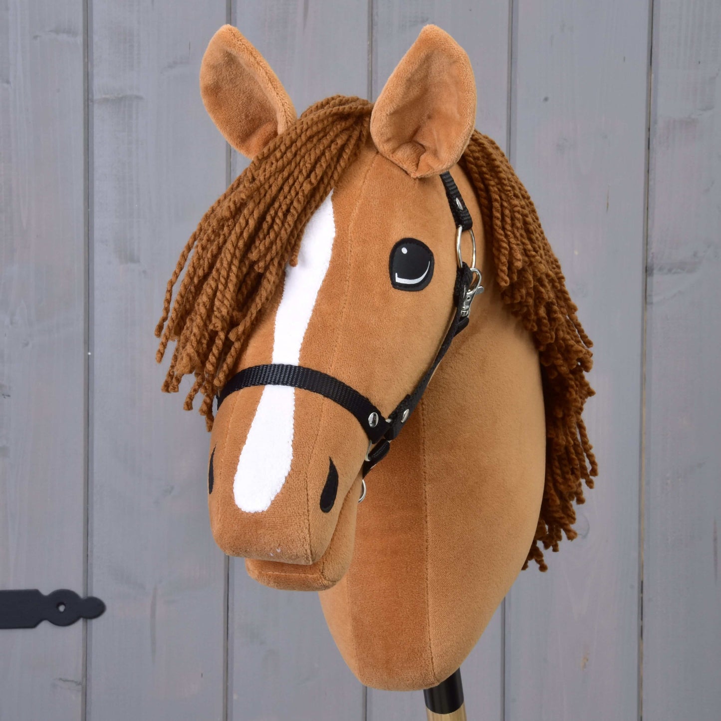 Hobby Horse Wendy avec licol noir de Hobby Horse LarDen, avec tête en peluche marron, crinière tressée en fil marron, marque blanche sur le museau et licol noir, inspiré d’une jument alezane et présenté sur fond en bois gris.