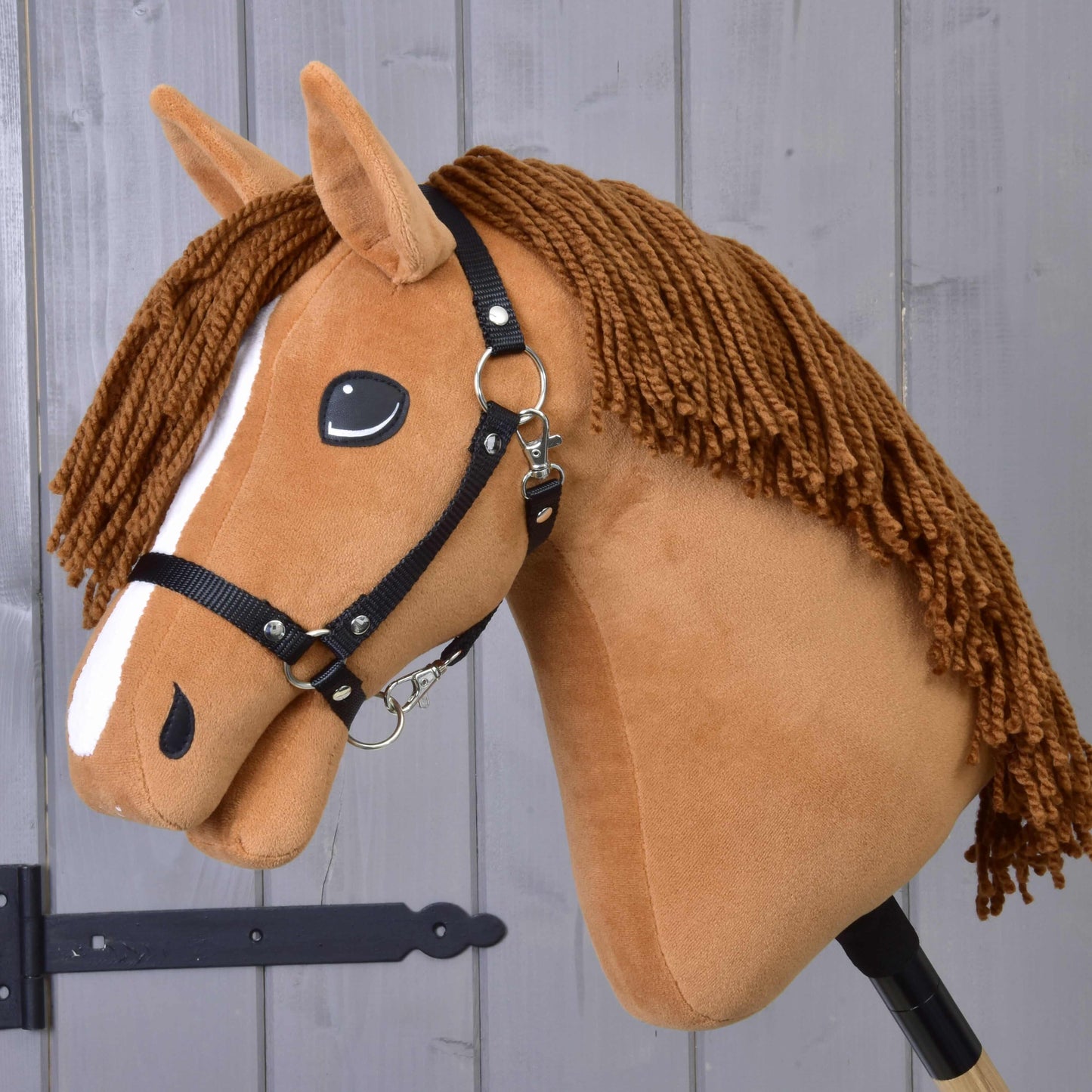 Hobby Horse Wendy avec licol noir de Hobby Horse LarDen, jouet en peluche jument alezane avec crinière en fil marron, liste blanche sur le visage et licol noir — idéal pour le jeu imaginatif — présenté sur fond de panneaux en bois.