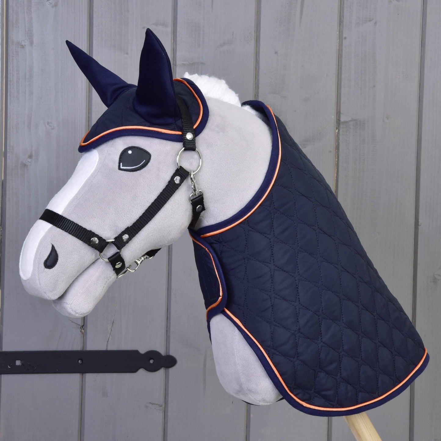 Hobby Horse LarDen „Set Winner“ zeigt einen Stoffpferdekopf auf einem Stock, gekleidet in eine gesteppte dunkelblaue Decke und passende Fliegenhaube mit orangefarbenem Besatz, vor einer Holzwand.