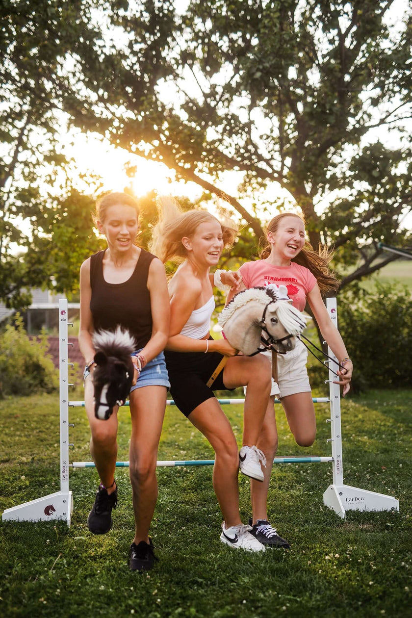 Drei junge Frauen lachen, während sie draußen laufen und über eine Hürde springen, jede mit einem Hobby Horse in der Hand, während die Sonne durch die Bäume hinter ihnen scheint und sie den Spaß am Hobby Horsing genießen.