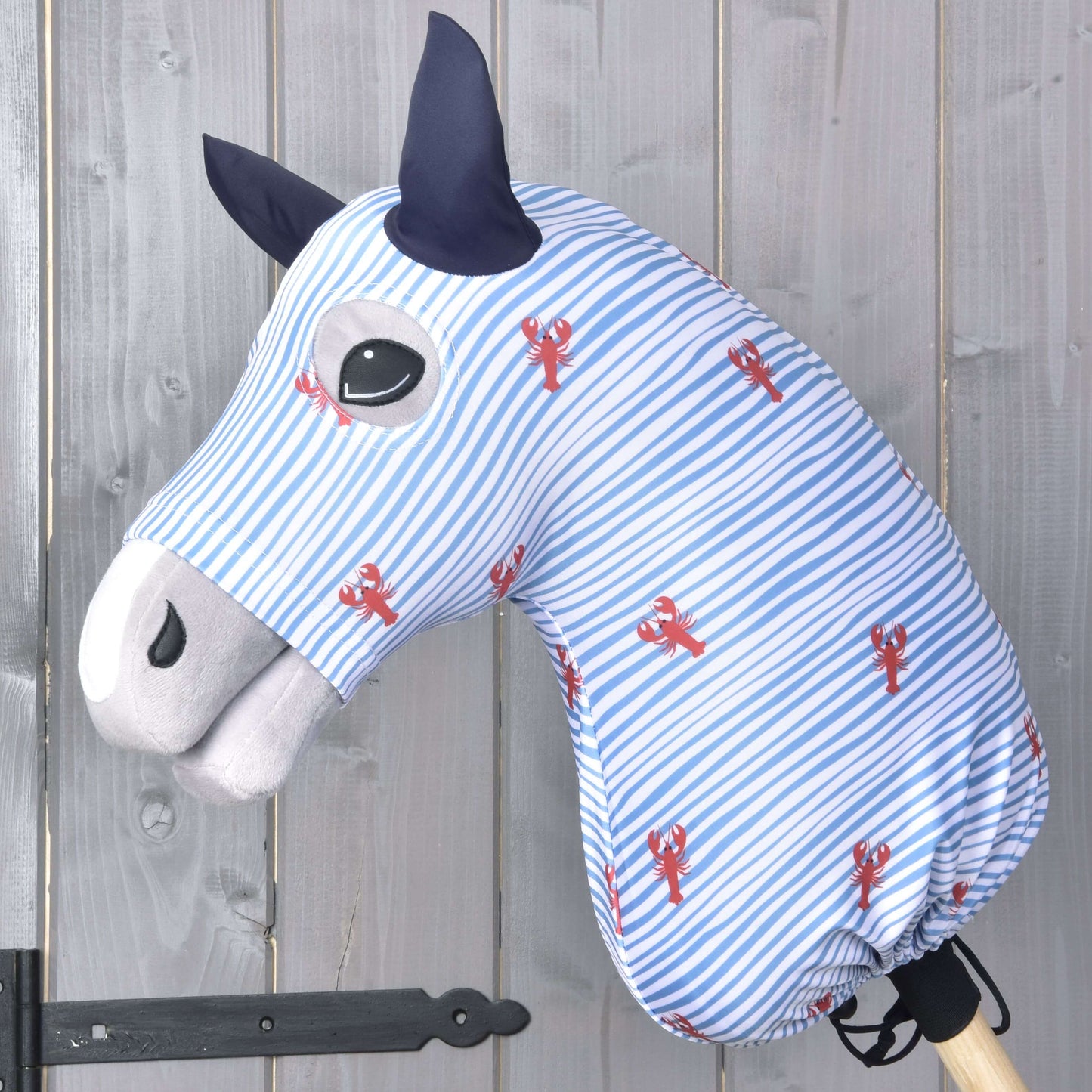 Hobby Horse Schutzanzug Andromeda (Lycra) von Hobby Horse LarDen in blau-weißen Streifen mit roten Hummermotiven bedeckt das Spielzeugpferd, das schwarze Augen, dunkelblaue Ohren und eine hellgraue Schnauze vor einer Holzwand zeigt.