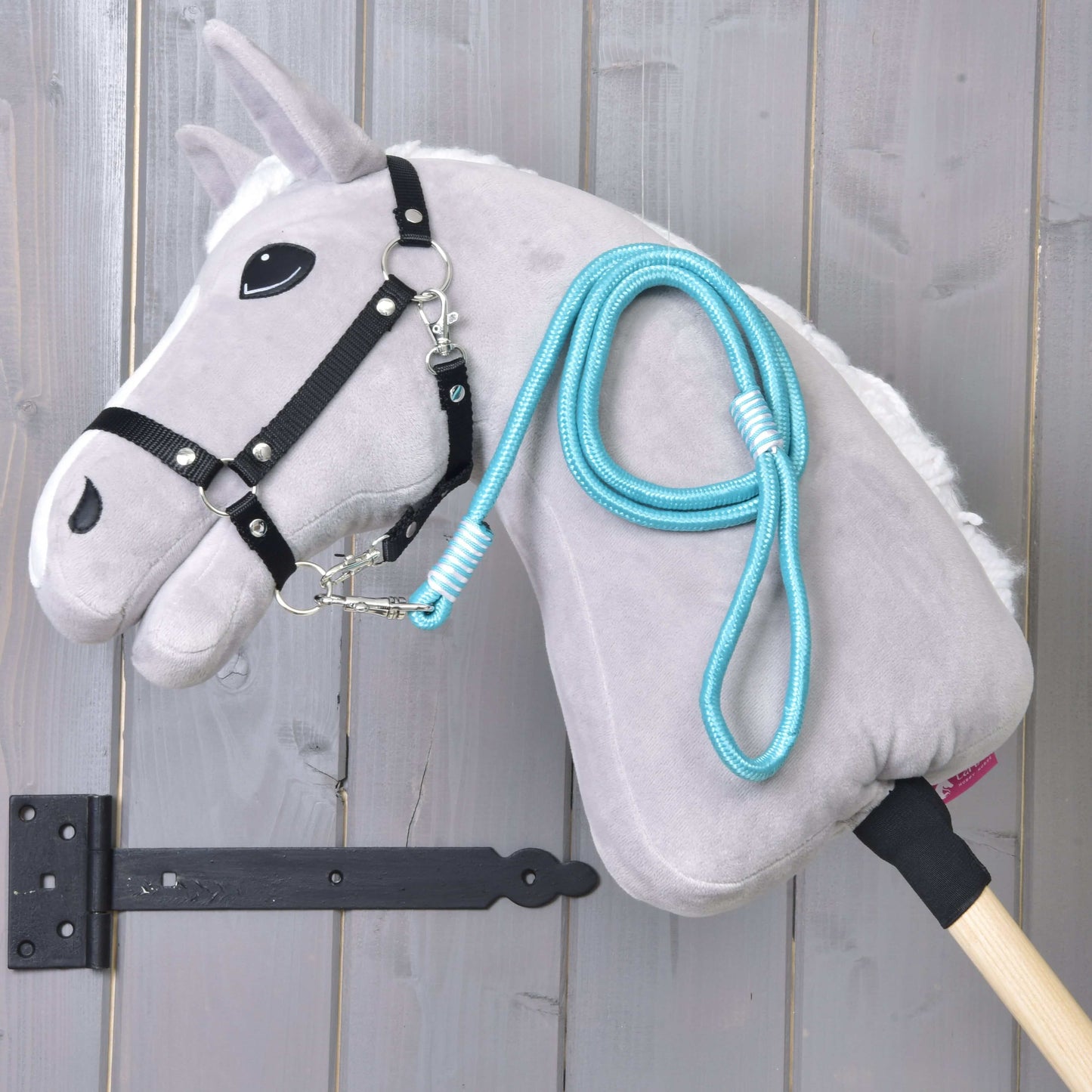 Hobby Horse - Führstrick Atlantis von Hobby Horse LarDen mit grauem Plüschkopf, schwarzem Zaumzeug für Hobby Horse, blauem Führstrick und Holzstab vor senkrechten Holzplanken