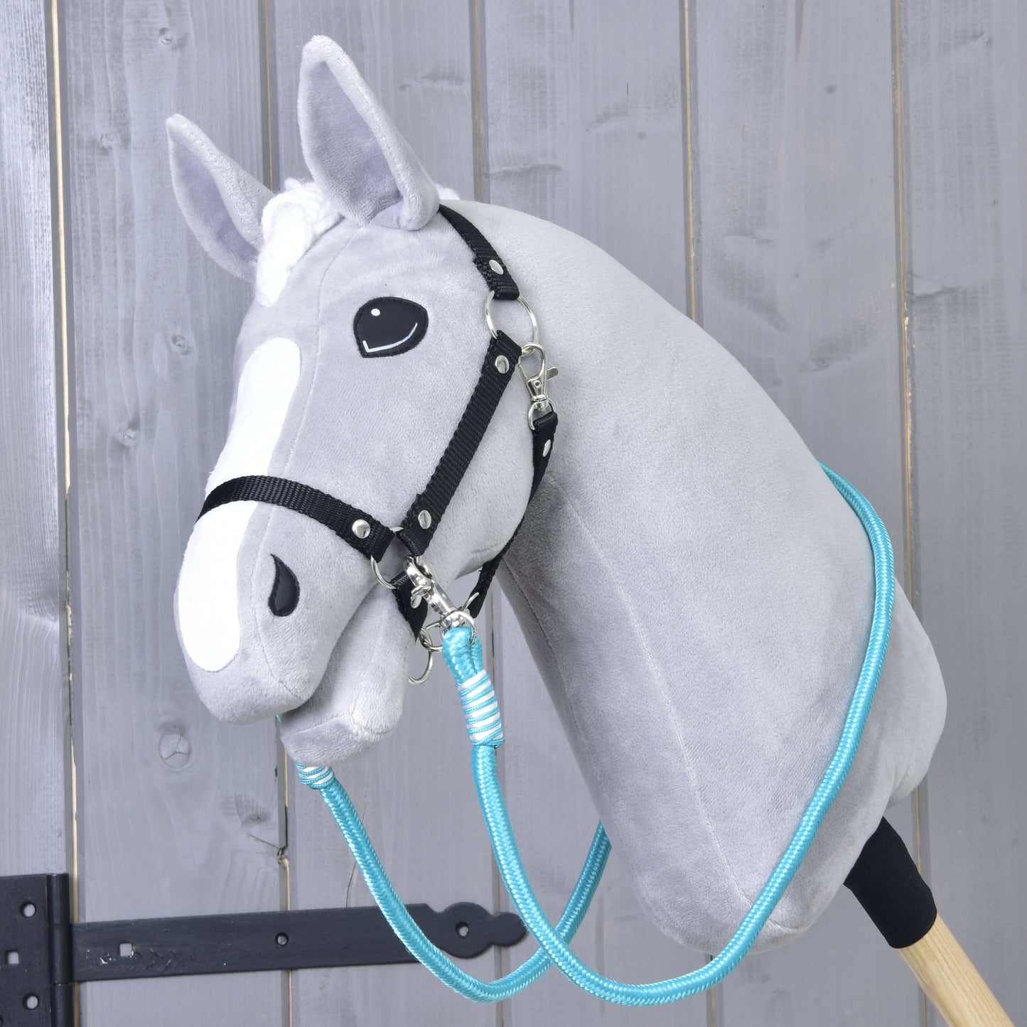 Rênes Atlantis de Hobby Horse LarDen sur un Hobby Horse gris en peluche avec bride noire et rênes bleues en corde de polypropylène, monté sur un bâton en bois devant un mur rustique en bois.