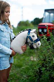 Lifestyle photo: a girl in a denim jacket holds the Hobby Horse Aurora with Black Halter by Hobby Horse LarDen beside a hedge, a red tractor blurred in the background - cream-white plush head, black and white striped yarn mane - shown here with the fur-lined LarDen Blue halter.
