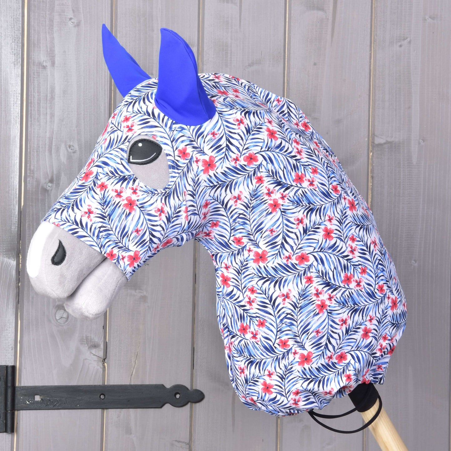 La Combinaison de protection Hobby Horse Avatar (Lycra) de Hobby Horse LarDen présente une tête en tissu à motif floral bleu et rouge, des oreilles en feutrine bleue, une crinière grise, et comprend une housse lavable. Elle est montée sur un bâton en bois devant un fond en panneaux de bois.