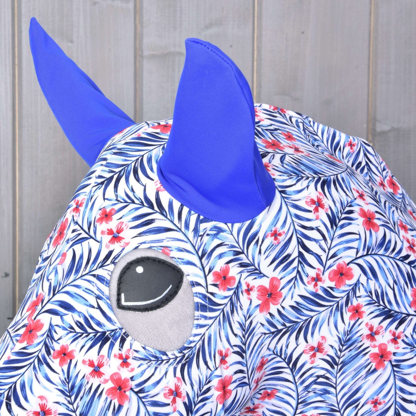 Gros plan sur la combinaison de protection « Avatar » (Lycra) de Hobby Horse LarDen montrant des oreilles bleues, un visage avec feuilles bleues et fleurs rouges, et un grand œil gris, le tout sur fond en bois. Le tissu lavable convient aux Hobby Horses.