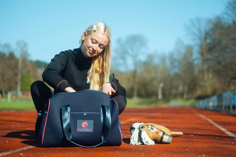 Jeune femme aux longs cheveux blonds assise sur une piste, regardant dans un sac de sport bleu marine marqué LarDen. Un hobby horse est posé au sol à côté d'elle, suggérant sa passion pour le Hobby Horsing. Des arbres et les lignes de la piste sont visibles en arrière-plan.
