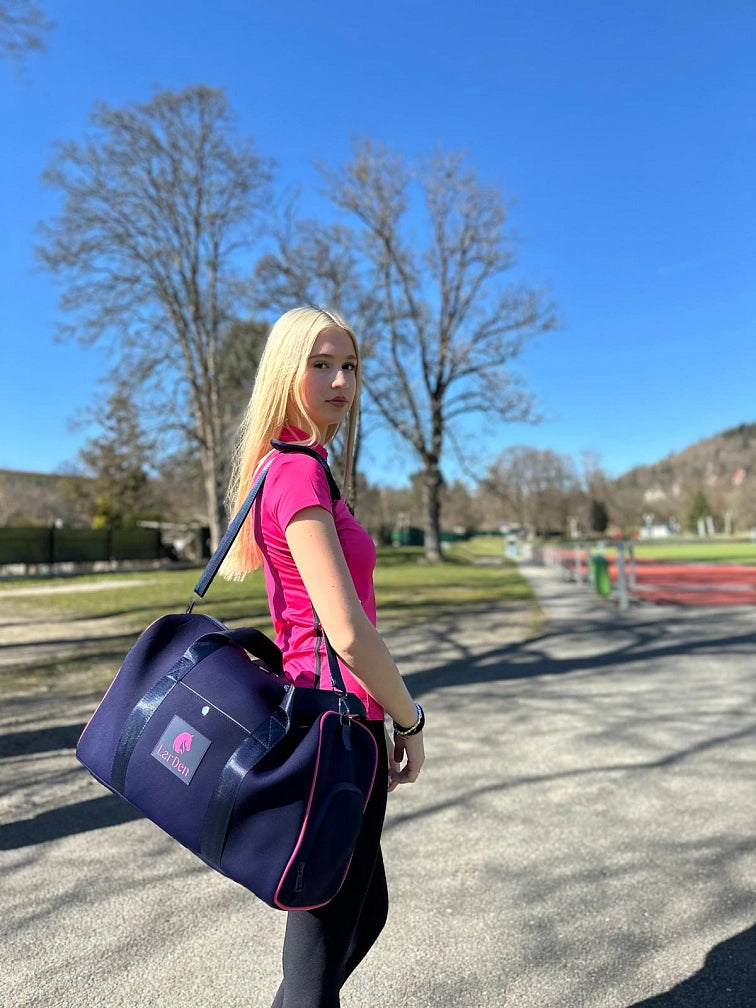 Jeune femme aux longs cheveux blonds portant un t-shirt de sport rose et un sac de sport bleu marine, debout dehors par une journée ensoleillée avec son Hobby Horse, prête pour une journée amusante de Hobby Horsing sous un ciel bleu clair et des arbres feuillus.