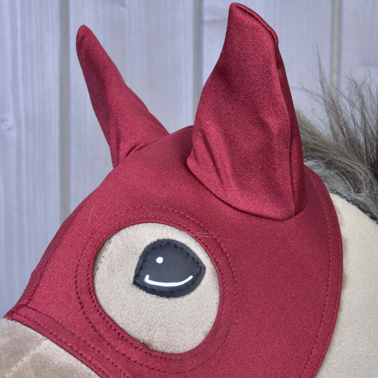 Gros plan sur une peluche Hobby Horse LarDen portant le masque de cheval Rouge Étable, avec des oreilles pointues et une crinière grise, sur fond en bois clair.