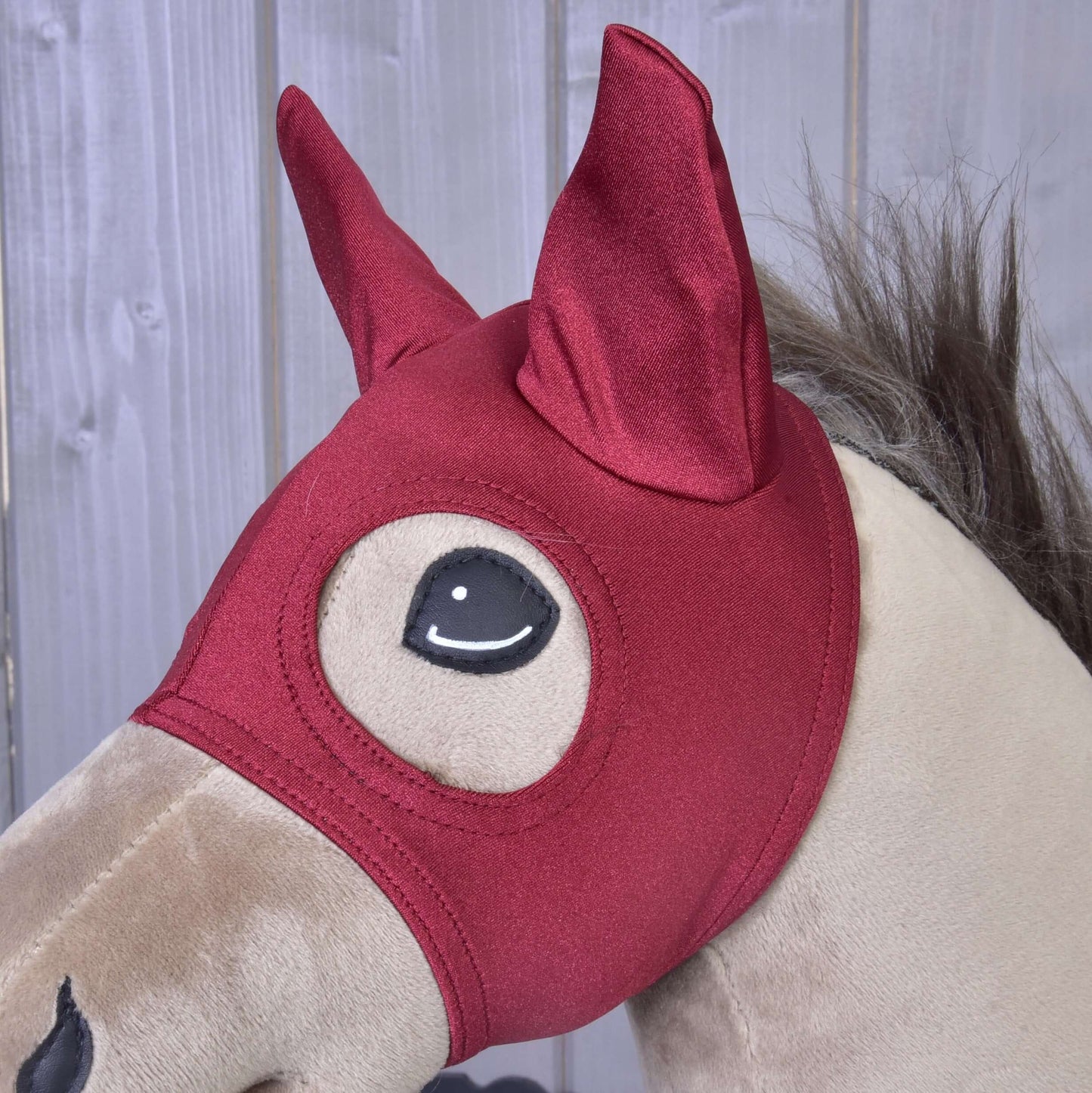 Un Hobby Horse en peluche beige de Hobby Horse LarDen porte le masque de cheval Rouge Étable, avec des oreilles pointues et de grandes ouvertures pour les yeux de style dessin animé, sur fond de panneaux en bois.