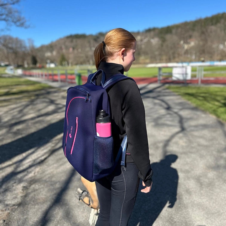 Jeune femme aux cheveux roux en queue de cheval marchant dehors par une journée ensoleillée, portant des vêtements de sport noirs et un sac à dos bleu marine avec une gourde rose. Des arbres et une piste d’athlétisme en arrière-plan suggèrent qu’elle se rend à son activité de Hobby Horsing préférée ou en revient.