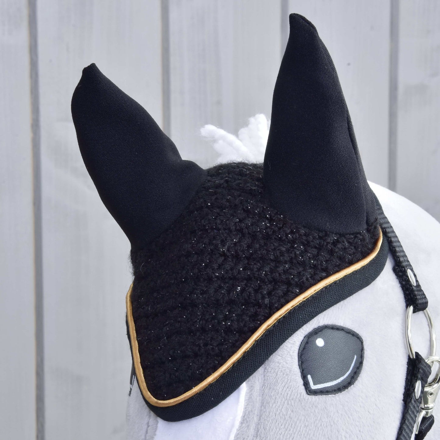 Gros plan sur un cheval en peluche de Hobby Horse LarDen portant le Bonnet d’oreilles au crochet Noir Or pour la protection contre les insectes. Le cheval a une bride, une crinière blanche et se trouve devant un mur en bois clair.