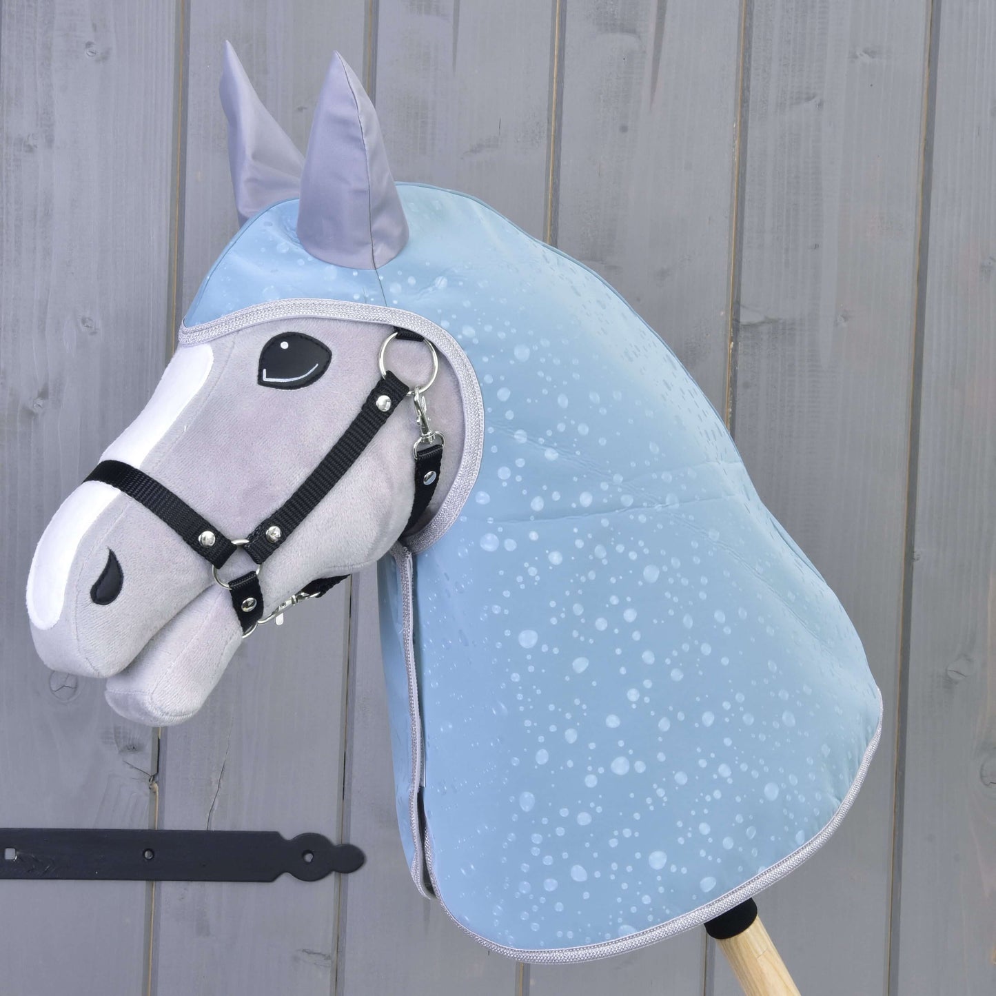 Imperméable Gouttes Bleues de Hobby Horse LarDen, présenté sur un Hobby Horse gris et blanc avec bride noire, exposé devant une clôture en bois clair, un accessoire idéal par tous les temps pour votre Hobby Horse.
