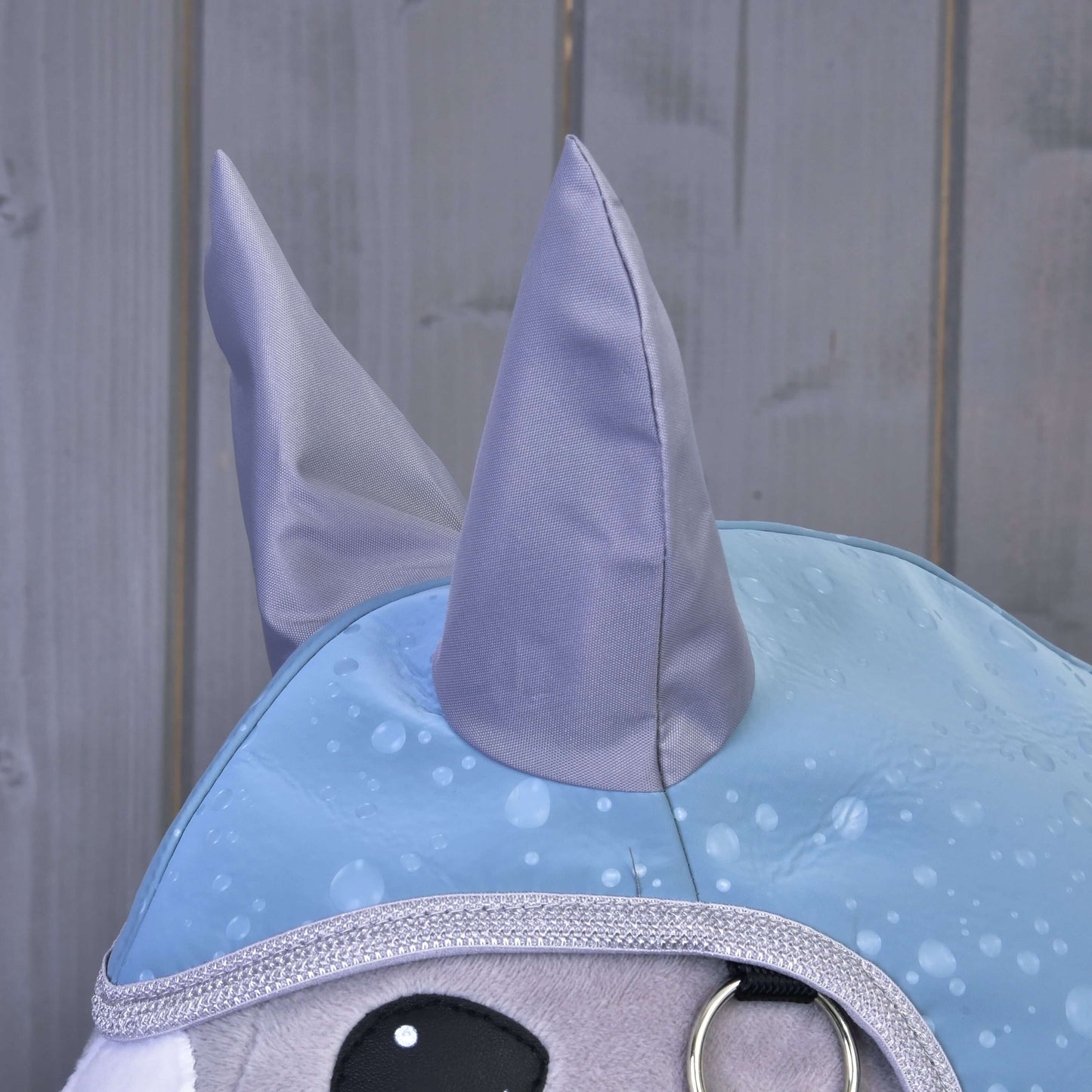 Gros plan sur une peluche Hobby Horse LarDen en tenue « Raincoat Blue Drops », avec oreilles grises brillantes, bordure argentée, grands yeux noirs et manteau bleu à motif gouttes de pluie. Des panneaux en bois gris lisse forment l'arrière-plan.