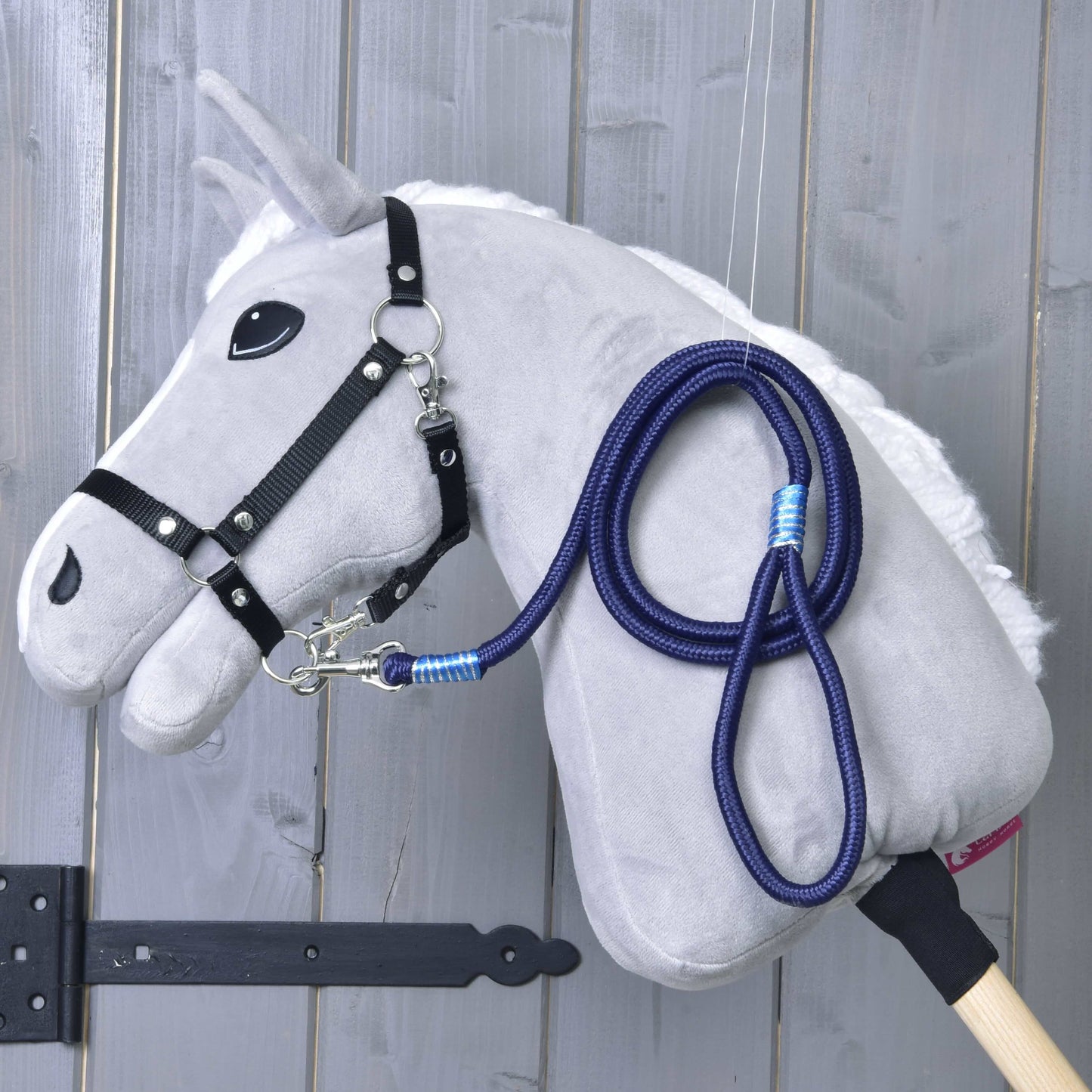The Hobby Horse LarDen "Hobby Horse - rope stripe Blues" features a plush gray horse with a black bridle and blue leash on a wooden stick, displayed against a gray wooden wall.