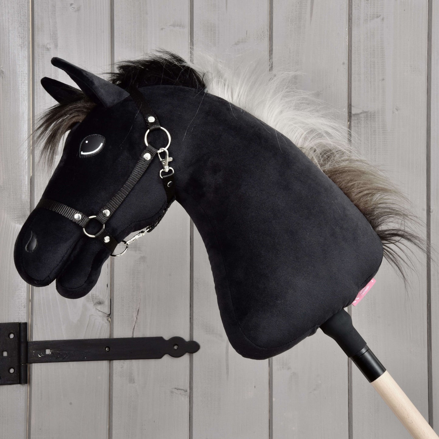 Hobby Horse Bond mit Schwarzem Halfter von Hobby Horse LarDen, ein schwarzes Hobby Horse aus Plüsch mit weißer Mähne, schwarzem Halfter und Zügeln an einem Holzstock, vor einer hellen Holzwand – perfekt für jeden Hobby Horse Bond-Fan.