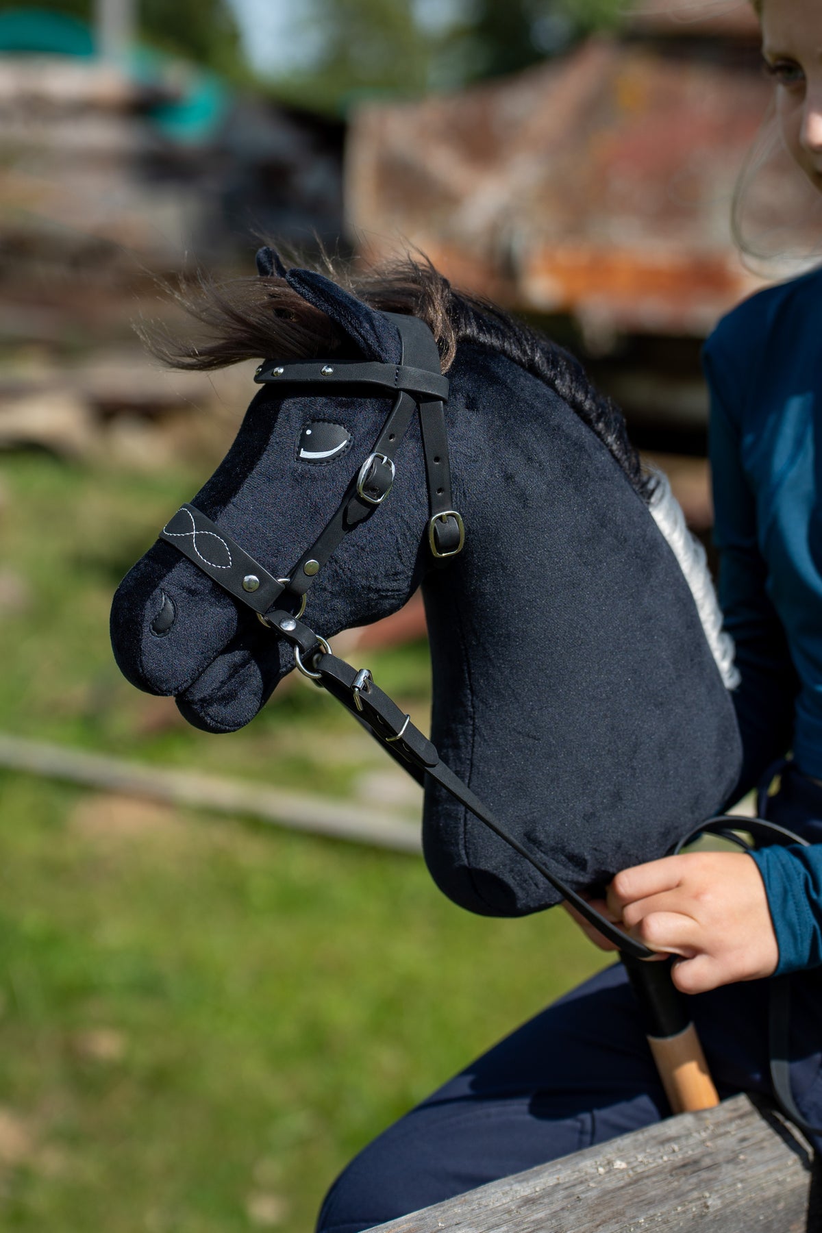 Kopf des Hobby Horse Bond mit schwarzem Halfter von Hobby Horse LarDen vor einem unscharfen Bauernhof – schwarzer Plüsch, dunkelbraune Garnmähne, schwarze Riemen mit Silberschnallen – hier mit dem verzierten gebisslosen Zaumzeug