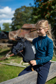 Lifestyle photo: a blonde girl in a teal top sits on a wooden rail in a sunny farmyard holding the Hobby Horse Bond with Black Halter by Hobby Horse LarDen - black plush head, dark brown yarn mane - shown here with the decorated bitless bridle.