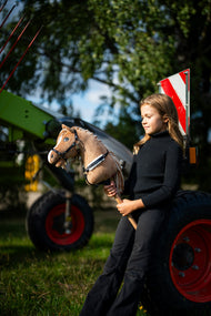 Lifestyle photo: a girl in a black turtleneck stands beside green farm machinery holding the Hobby Horse Bren with Black Halter by Hobby Horse LarDen - sandy-brown plush head, cream yarn mane - shown here with the Espresso breastgirth with fur.