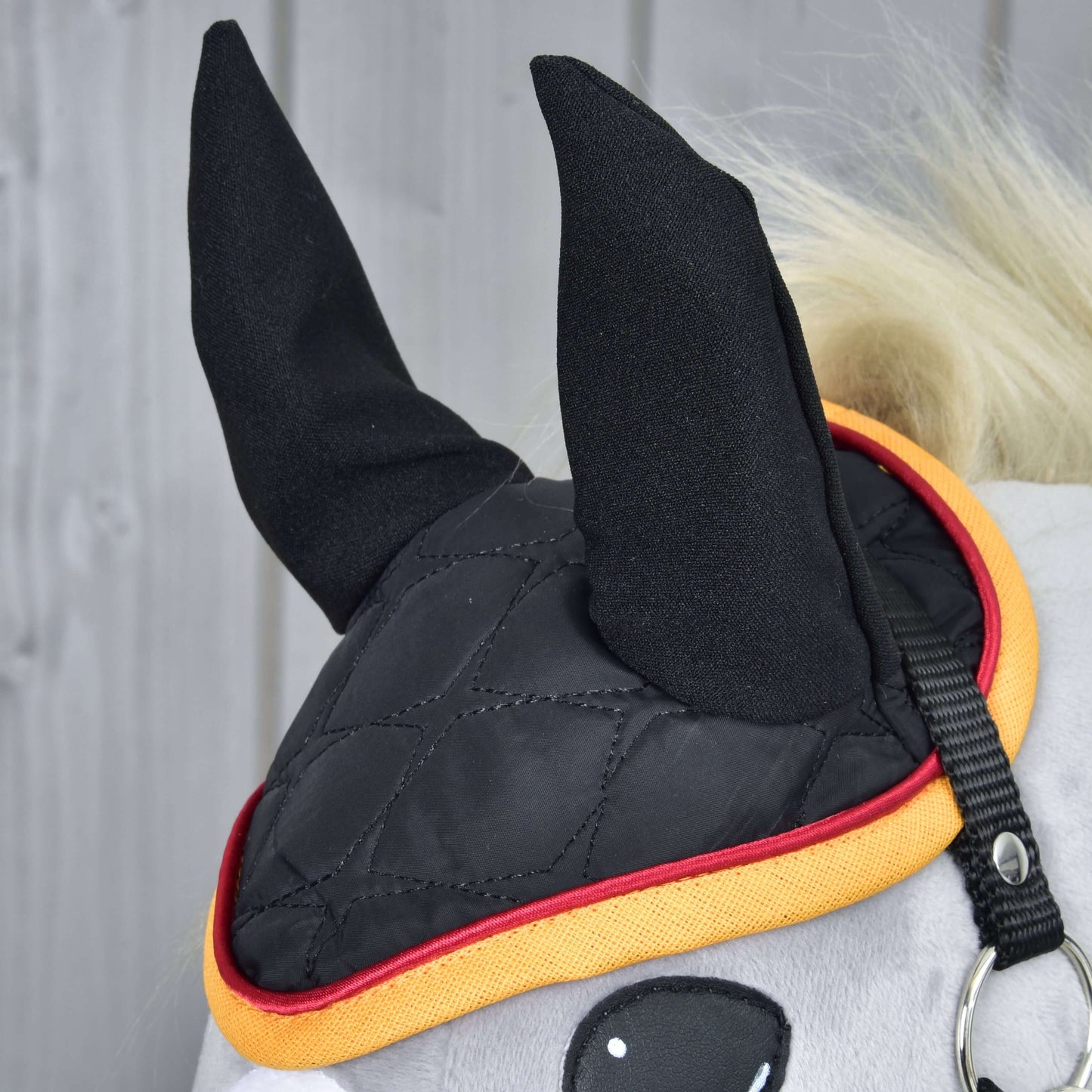 Gros plan sur un cheval portant le Bonnet d'oreilles Drapeau DE de Hobby Horse LarDen, un bonnet d’oreilles noir matelassé avec bordures jaune et rouge pour la protection contre les insectes, couvrant les oreilles et une partie de la tête sur fond en bois clair.