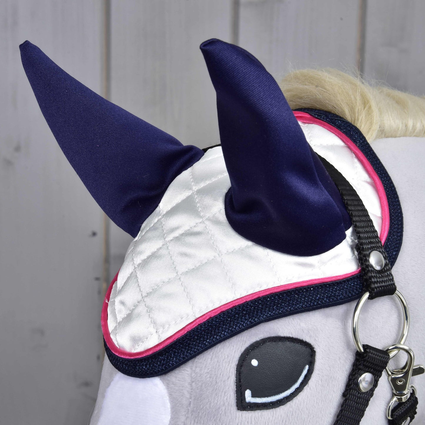 Gros plan sur la tête d'un Hobby Horse LarDen avec le Bonnet d'oreilles Numéro 1 — blanc matelassé avec oreilles bleu foncé et bordure rose — pour la protection contre les insectes, avec une bride noire, sur fond en bois clair.