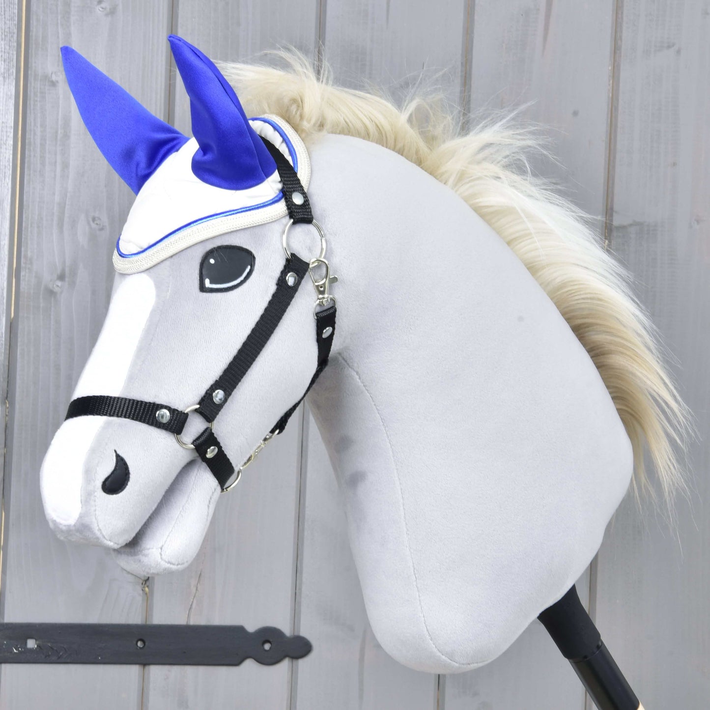 Das Hobby Horse LarDen zeigt einen plüschigen hellgrauen Kopf mit weißer Schnauze, schwarzem Zaumzeug und cremefarbener Mähne und trägt die Ohrenkappe Cream Pearl zum Insektenschutz. Es ist auf einem Stock montiert und vor Holz-Hintergrund dargestellt.