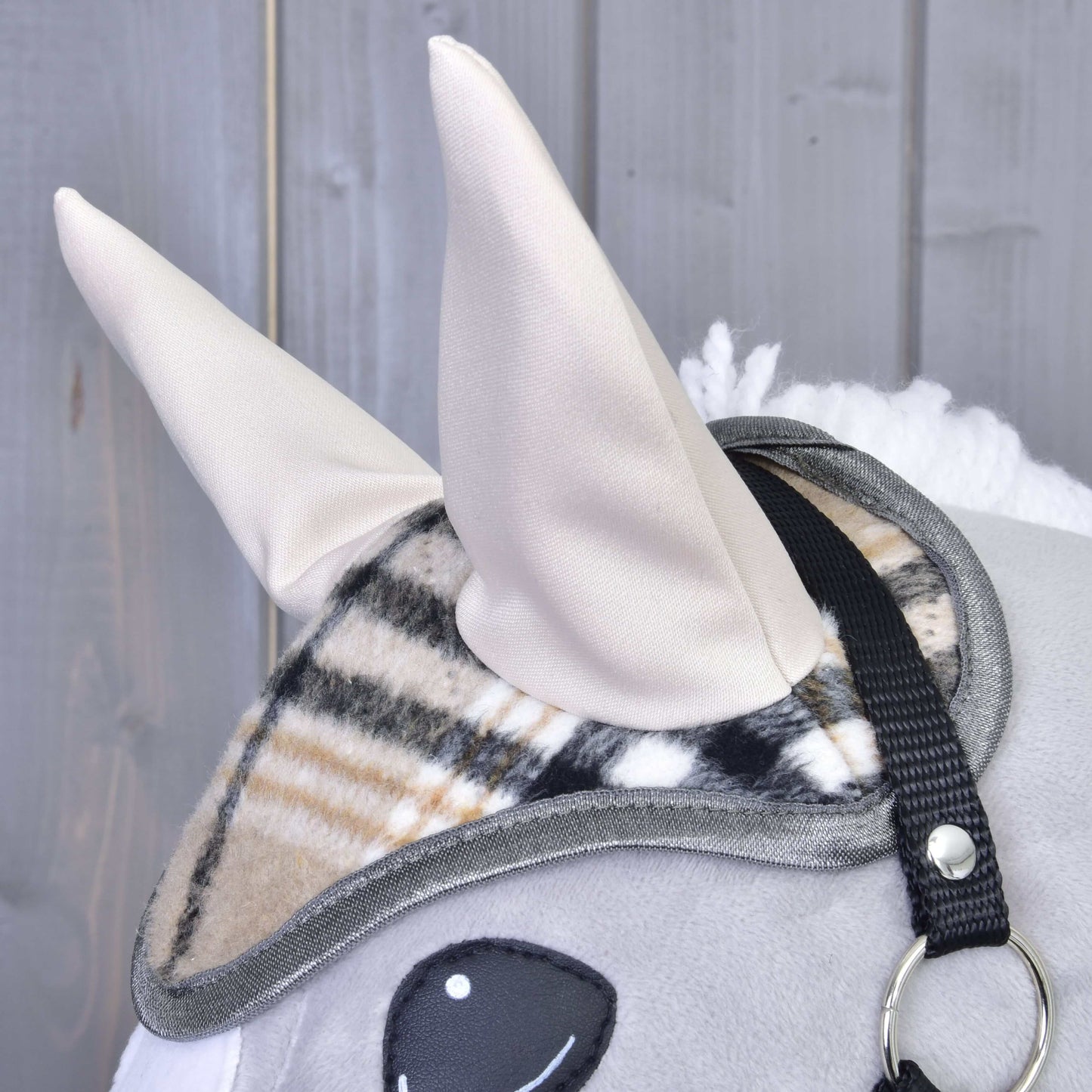 Gros plan sur un Hobby Horse en peluche de Hobby Horse LarDen avec deux oreilles en tissu beige, portant le Bonnet d’oreilles Cremo England à motif carreaux pour la protection contre les mouches, avec sangle de bride noire, sur fond en bois gris.
