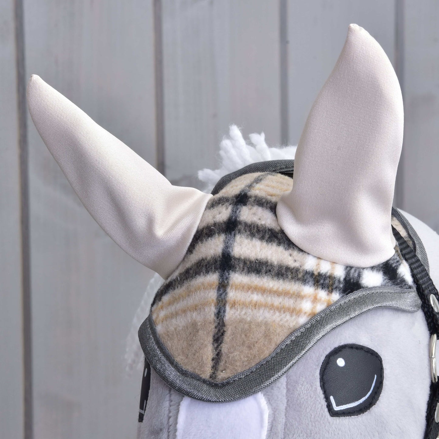 Gros plan sur le Bonnet d’oreilles Cremo England de Hobby Horse LarDen : oreilles douces de couleur crème, bride à carreaux pour la protection contre les mouches et crinière grise, sur fond en bois clair.