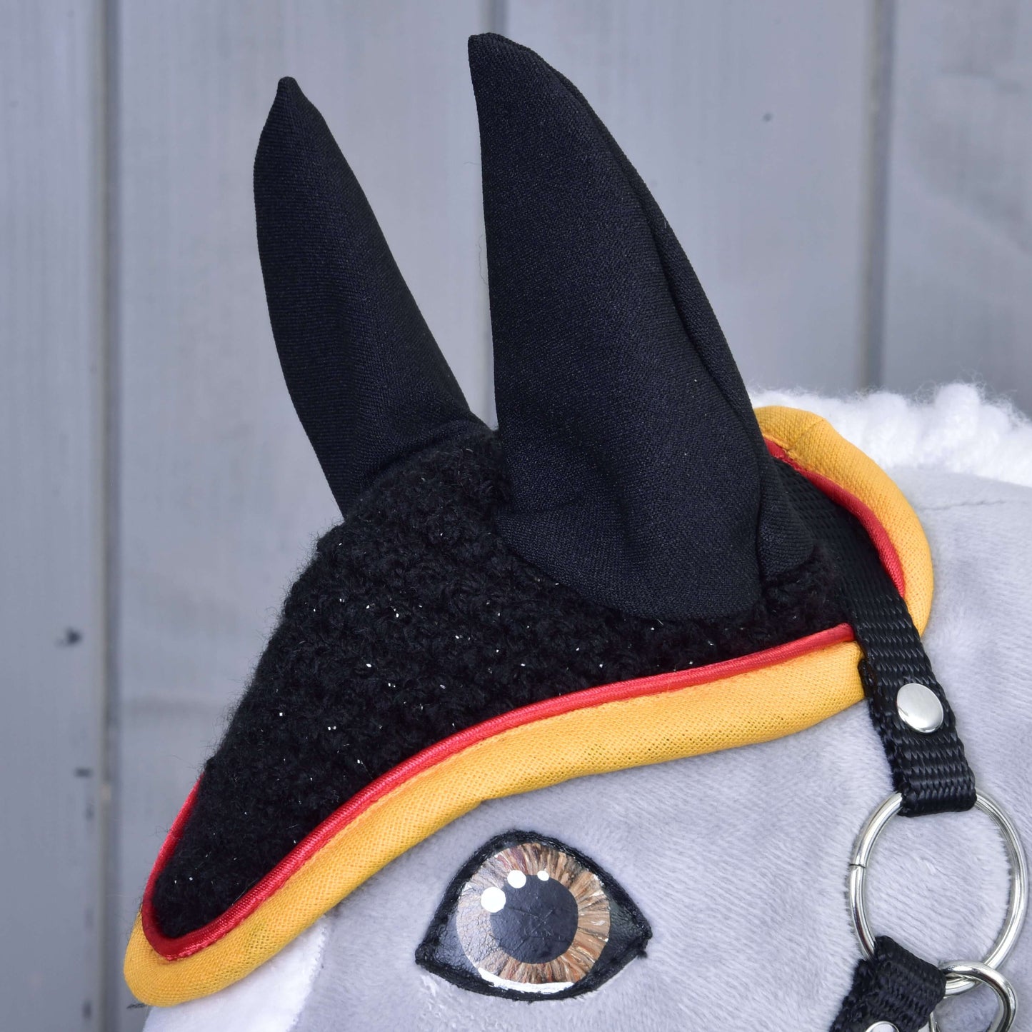 Nahaufnahme eines grauen Hobby Horse von Hobby Horse LarDen mit der Ohrenkappe gehäkelt Flag als Insektenschutz, mit gelber und roter Einfassung, einem glänzenden Auge, Metallring am Zaumzeug und weicher Mähne vor grauem Holzhintergrund.