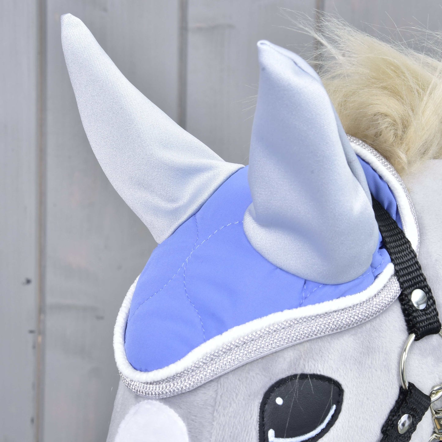 Gros plan sur un jouet Hobby Horse en peluche gris clair de Hobby Horse LarDen portant le Bonnet d'oreilles violet clair pour la protection contre les mouches, avec bordure brillante, sangles noires et œil blanc et noir, sur fond en bois.