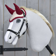 The Hobby Horse LarDen plush hobby horse with a white body, black bridle, and cream mane features the Ear Bonnet Maroon Star for insect protection, shown on a stick against a wooden background.
