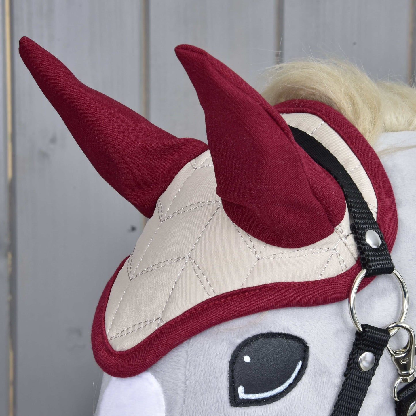 Gros plan sur un Hobby Horse LarDen avec le Bonnet d'oreilles Étoile Bordeaux, avec matelassage beige pour la protection contre les insectes, deux formes rouges dressées en forme de cornes et une bride noire. Des panneaux en bois clair apparaissent en arrière-plan.