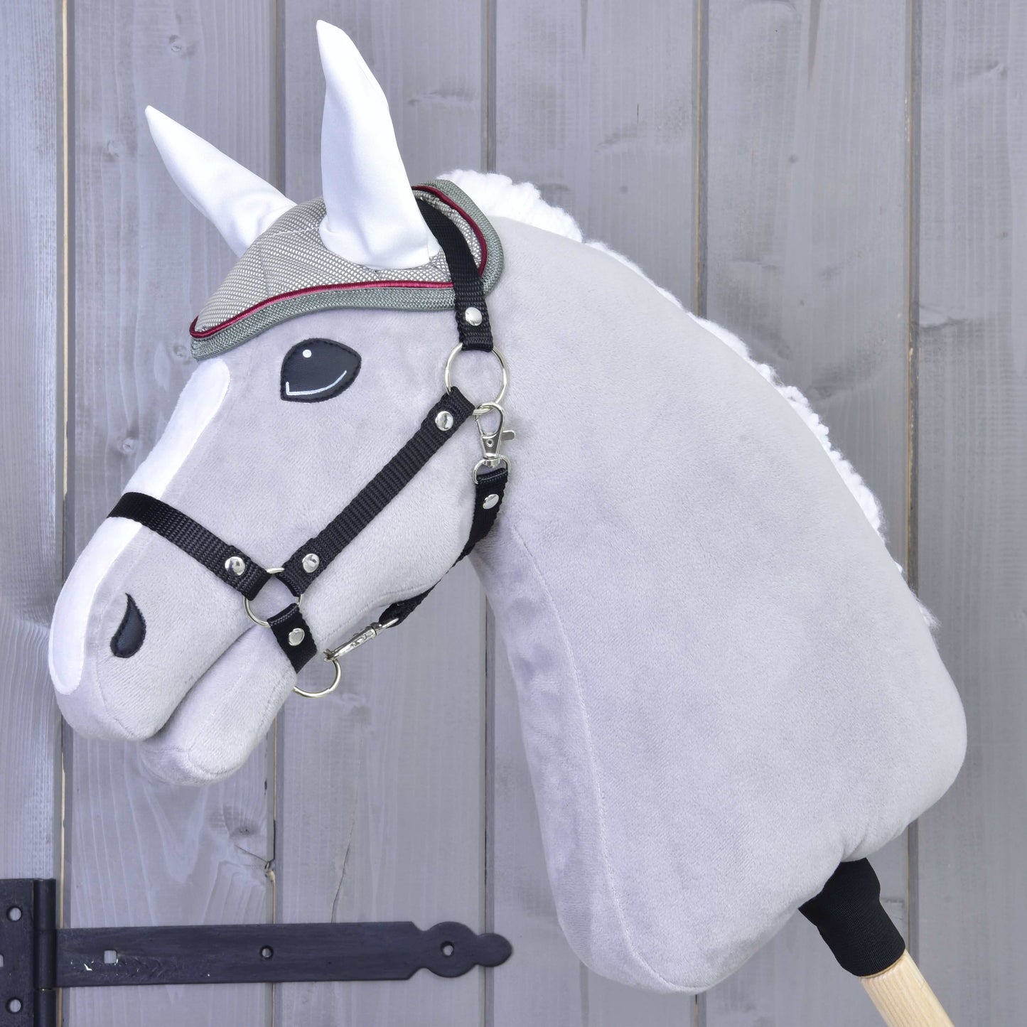 Gros plan sur un Hobby Horse en peluche gris de Hobby Horse LarDen, avec bride, frontal décoratif et Bonnet d'Oreilles Argent Élégance pour la protection contre les insectes, devant une clôture en bois.