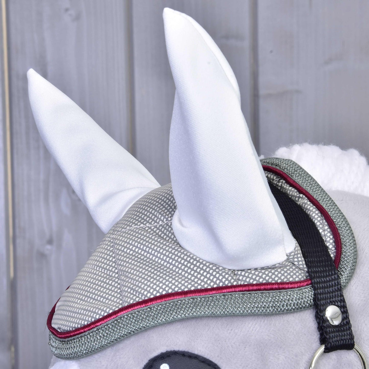 Gros plan sur un Hobby Horse gris de Hobby Horse LarDen portant le Bonnet d'Oreilles Argent Élégance et une bride grise et rouge, avec grandes oreilles blanches, sur fond de panneaux en bois.