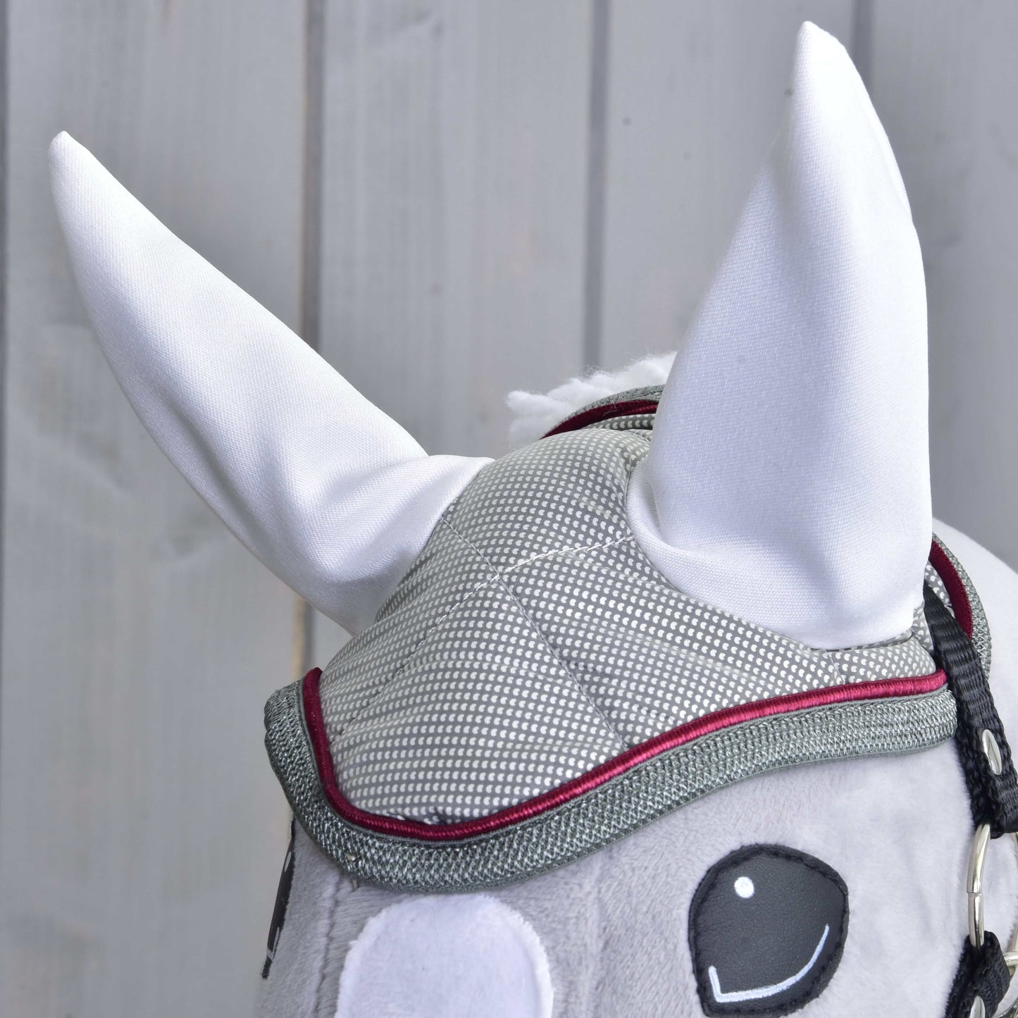 Gros plan sur le Bonnet d'Oreilles Argent Élégance de Hobby Horse LarDen : bonnet en maille grise avec bordure rouge, grandes oreilles blanches dressées et yeux noirs brodés, parfait pour la protection contre les insectes, sur fond en bois clair.
