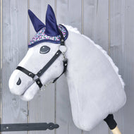 The Hobby Horse LarDen Ear Bonnet Flower is a plush hobby horse toy featuring a light gray head, black bridle, and sparkly purple and silver ear bonnet for insect protection, shown against a wooden background.